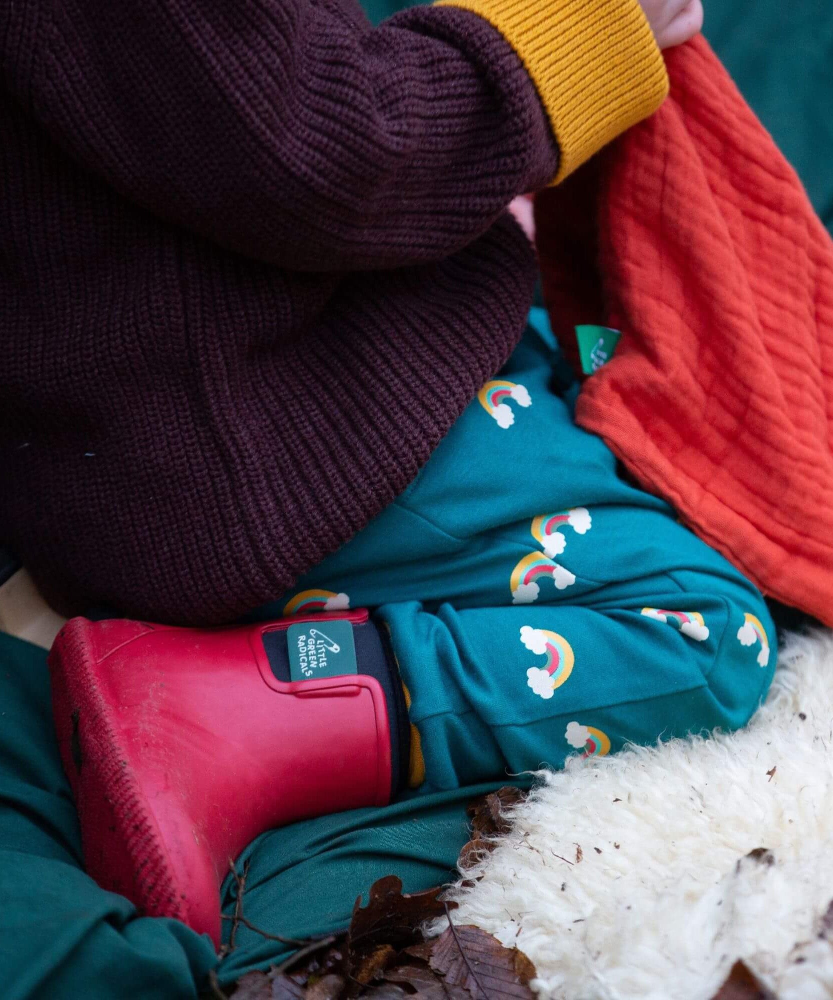 A close up of a child sitting down and wearing the Little Green Radicals turquoise rainbow joggers showing the print detail on organic cotton fabric.