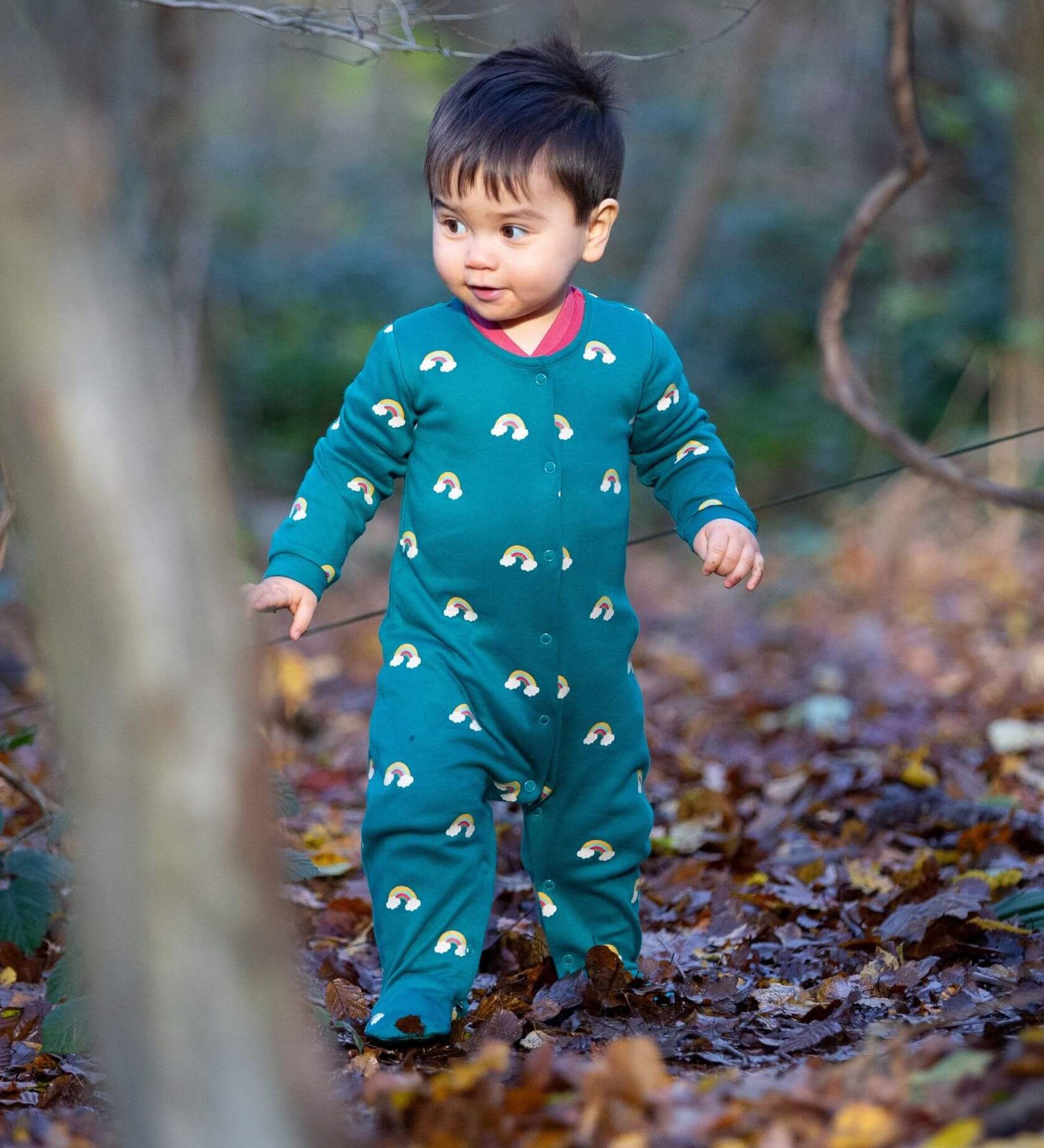 A child walking in the woods and wearing the Little Green radicals turquoise rainbow baby grow. A soft GOTS organic cotton all in one in turquoise with cute rainbow prints. Features popper fasteners on the front and around the crotch 