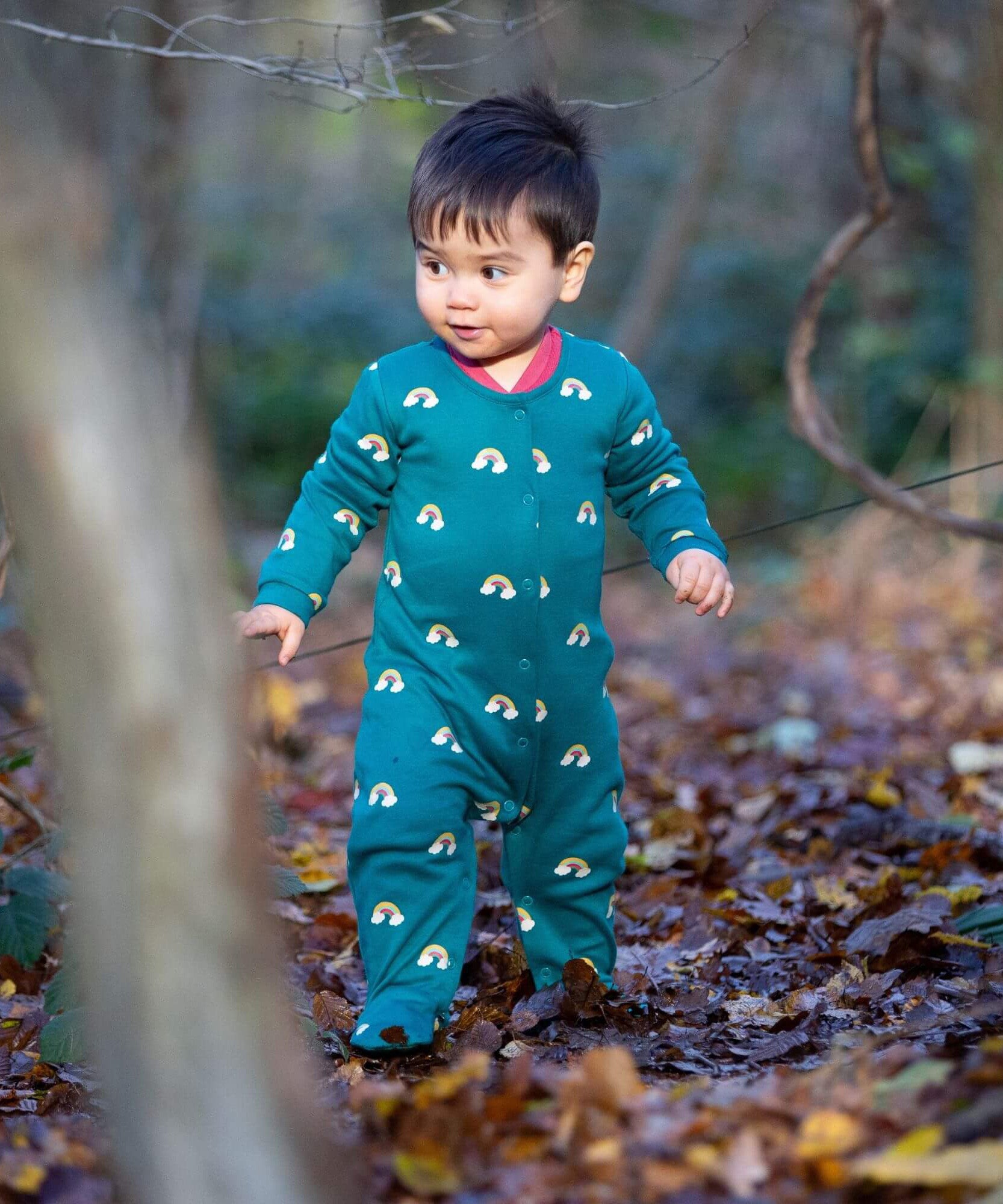 A child walking in the woods and wearing the Little Green radicals turquoise rainbow baby grow. A soft GOTS organic cotton all in one in turquoise with cute rainbow prints. Features popper fasteners on the front and around the crotch 