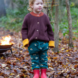 A child standing on leaves in a woodland wearing the Little Green Radicals comfy joggers in turquoise rainbow. A turquoise jogger with cute prints of rainbows on soft GOTS organic cotton fabric