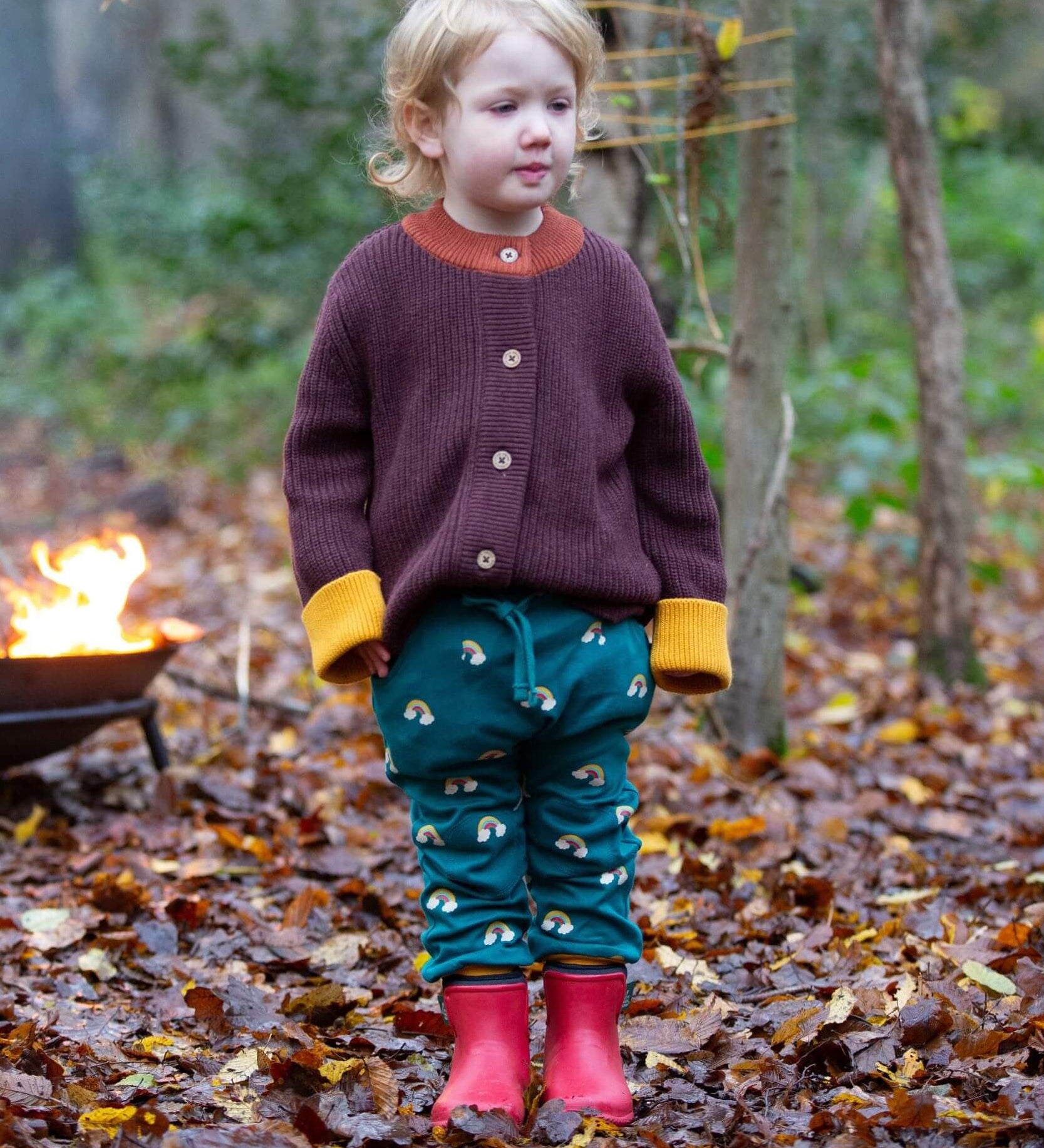 A child standing on leaves in a woodland wearing the Little Green Radicals comfy joggers in turquoise rainbow. A turquoise jogger with cute prints of rainbows on soft GOTS organic cotton fabric