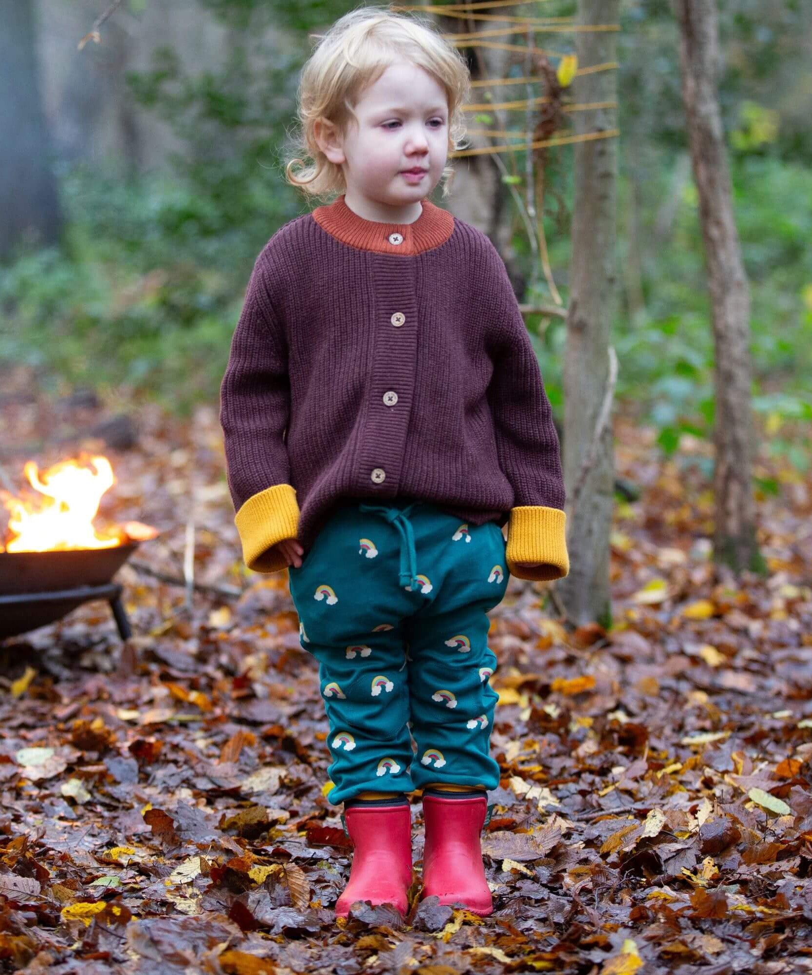 A child standing on leaves in a woodland wearing the Little Green Radicals comfy joggers in turquoise rainbow. A turquoise jogger with cute prints of rainbows on soft GOTS organic cotton fabric