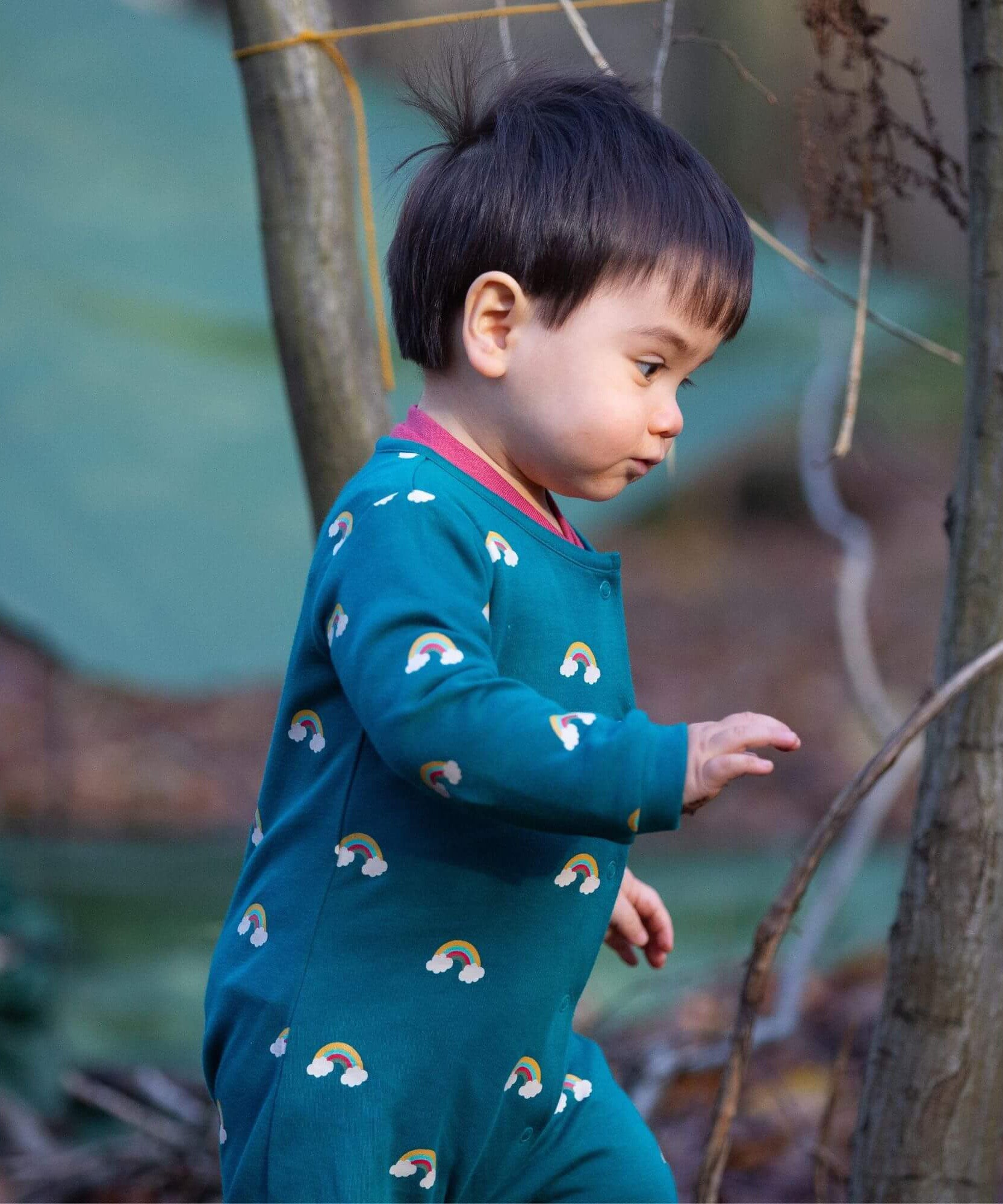 A child walking in the woods and wearing the Little Green radicals turquoise rainbow baby grow. A soft GOTS organic cotton all in one in turquoise with long sleeves and cute rainbow prints 