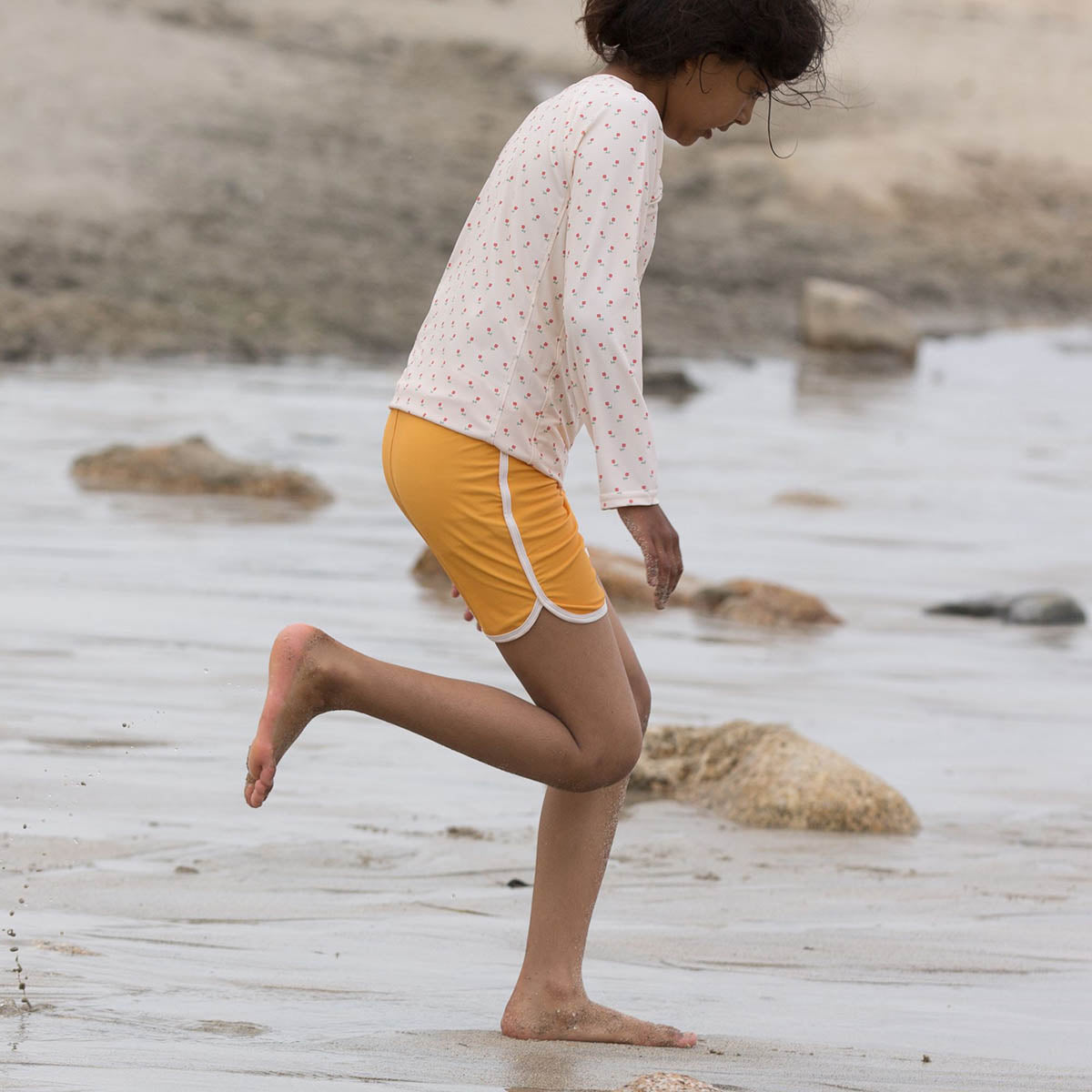 Girl running on sand wearing the LGR yellow swimming shorts and a rose print rash vest
