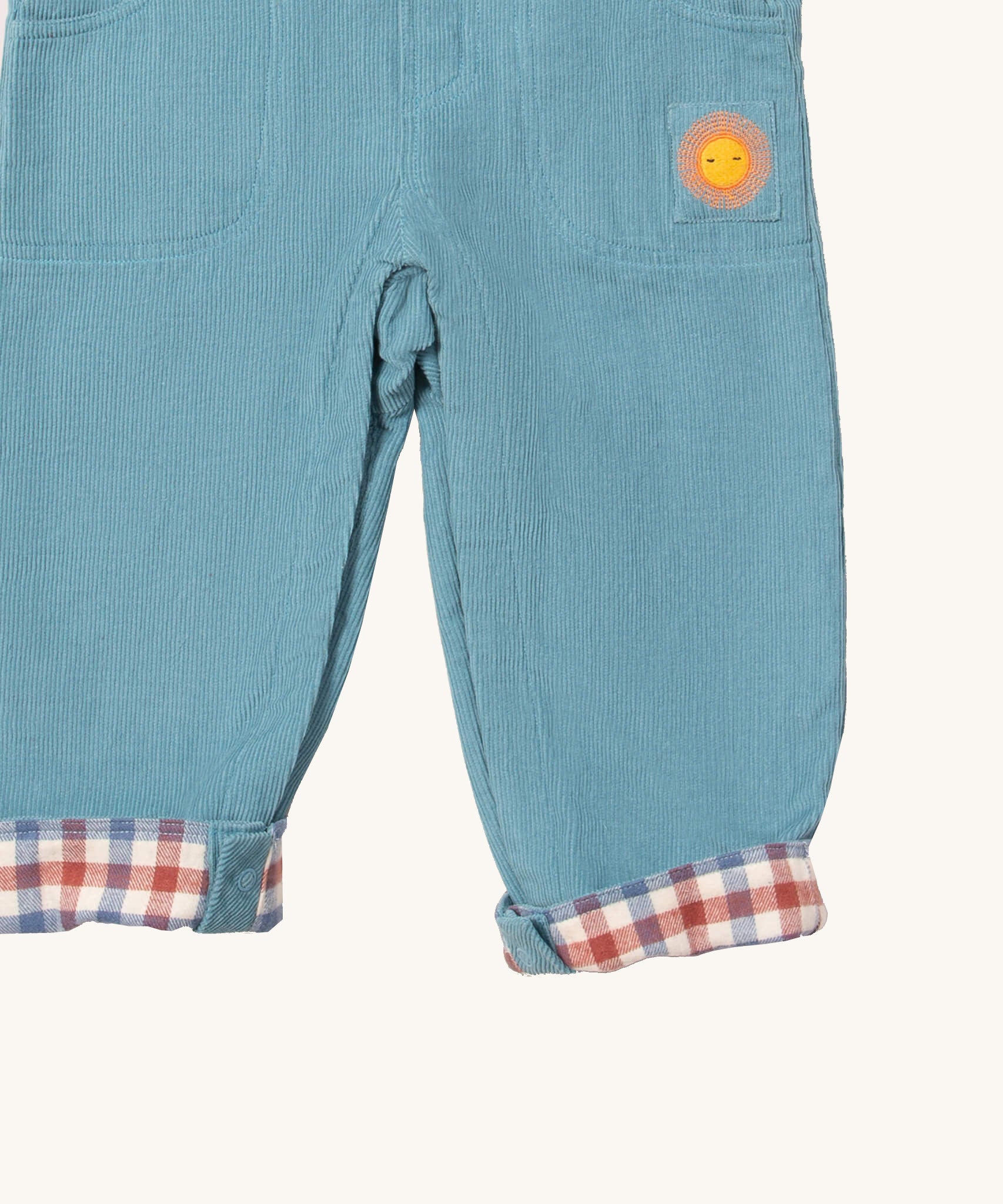 A close up of the Little Green Radicals pale blue lined corduroy dungarees showing the the sun embroidery under the side pocket and the turned up leg cuff showing the check design detail