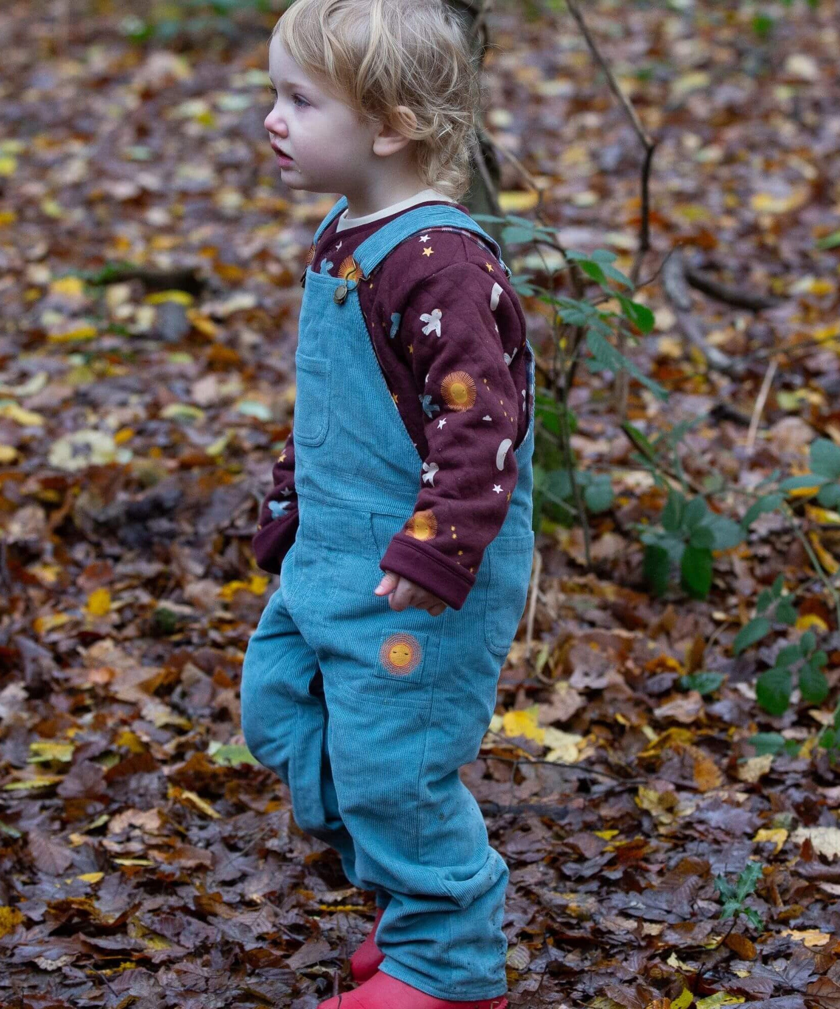 A child walking in the woods and wearing the pale blue Little Green Radicals corduroy dungarees over the quilted flying birds sweater
