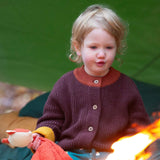 A child sitting outside and wearing the Little Green Radicals knitted cardigan in chocolate available at Babipur. A warm brown GOTS organic cotton cardigan with yellow arm cuffs, light brown collar and button fasteners. 