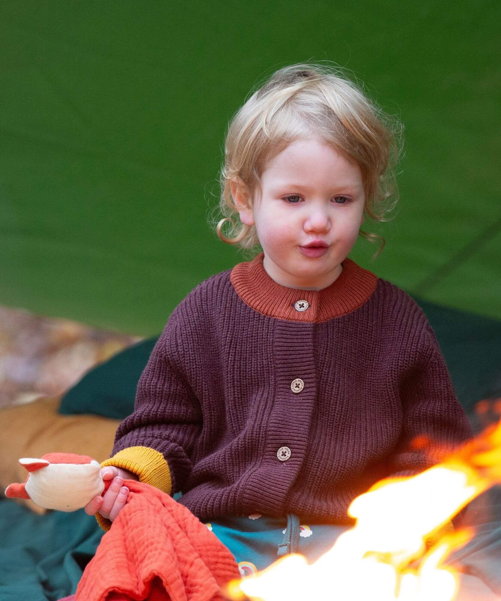 A child sitting outside and wearing the Little Green Radicals knitted cardigan in chocolate available at Babipur. A warm brown GOTS organic cotton cardigan with yellow arm cuffs, light brown collar and button fasteners. 