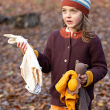 A child holding to a bunch of soft comforters outside and wearing the Little Green Radicals knitted cardigan in chocolate. A warm brown GOTS organic cotton cardigan with yellow arm cuffs and button fasteners. 