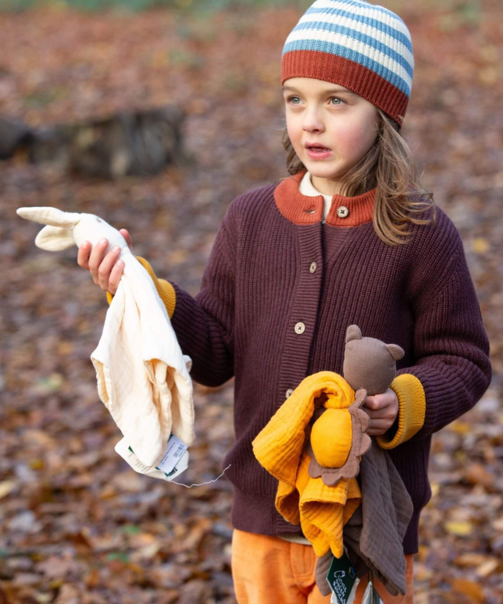 A child holding to a bunch of soft comforters outside and wearing the Little Green Radicals knitted cardigan in chocolate. A warm brown GOTS organic cotton cardigan with yellow arm cuffs and button fasteners. 