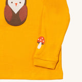 A close up of the Little Green Radicals gold brown owl applique long sleeve top showing the mushroom applique detail on dark yellow organic cotton fabric.