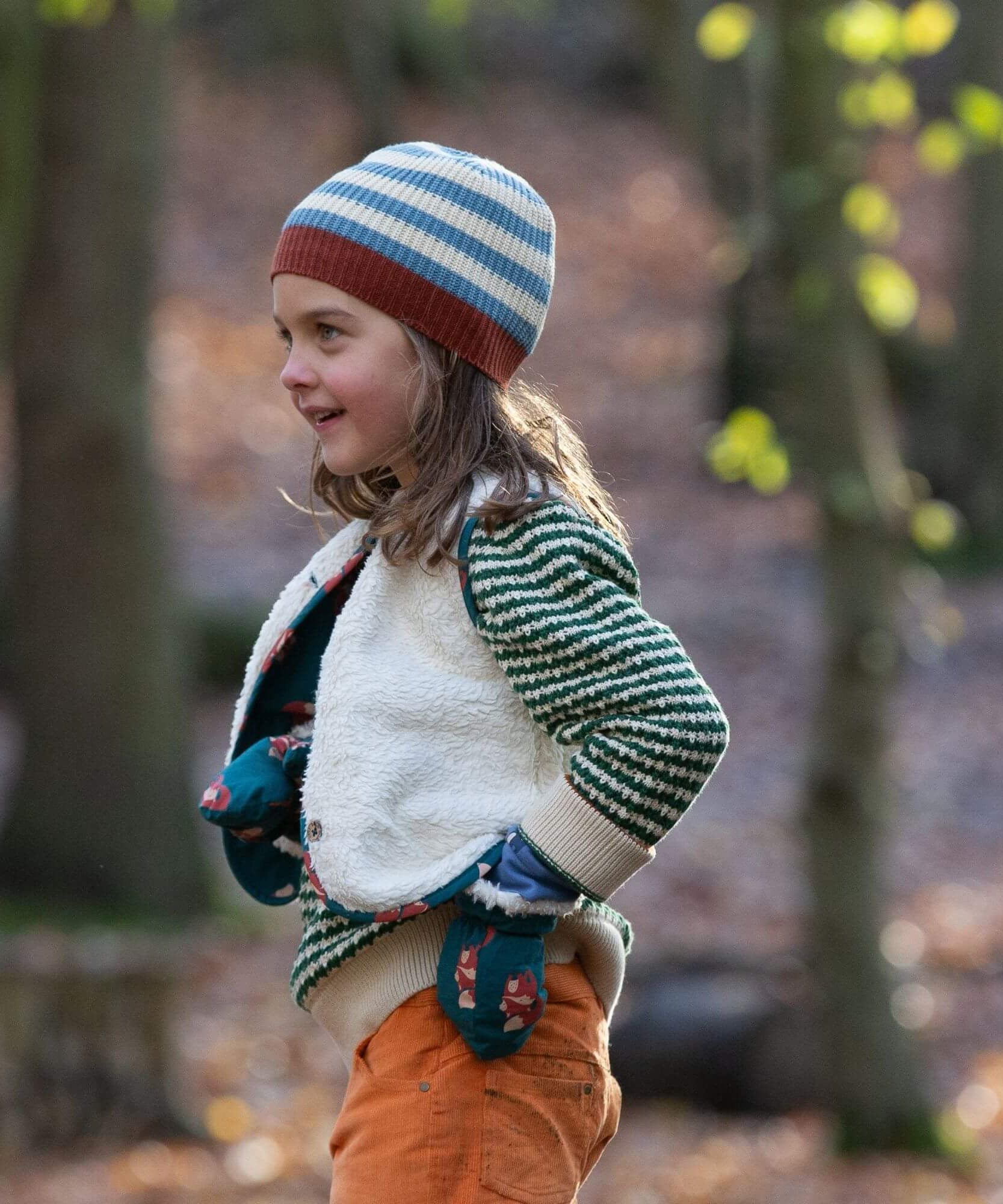 A child wearing the Little Green Radicals snuggly knitted jumper in olive stripe  with a reversible sherpa fleece fox gilet on top making a warm outfit available at Babipur.