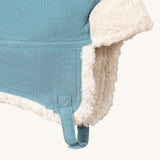 A close up of the Little Green Radials sherpa fleece cosy hat in pale blue. Showing the foldable ear flaps, the white sherpa fleece inner and matching corduroy chin strap on blue organic cotton fabric