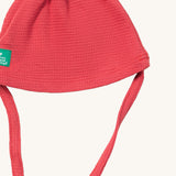 A close up of the Little Green Radicals pink berry waffle tie hat showing the tie strap for a secure fit and waffle knit detail on pink organic cotton fabric