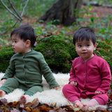 2 children wearing the holly berry pink and green chive Little Green Radicals waffle playsuit outside in woodland. A long sleeved, footless all in one suit made from GOTS organic cotton fabric with button fasteners and a small pouch chest pocket