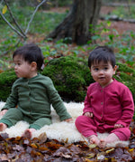 2 children wearing the holly berry pink and green chive Little Green Radicals waffle playsuit outside in woodland. A long sleeved, footless all in one suit made from GOTS organic cotton fabric with button fasteners and a small pouch chest pocket