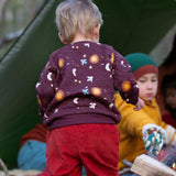 A child in woodland and wearing the Little Green Radicals flying birds quilted sweatshirt that's available at Babipur. A thick sweatshirt with quilted indents and cute prints of birds flying through a starry night on dark maroon organic cotton fabric.