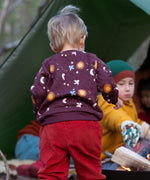 A child in woodland and wearing the Little Green Radicals flying birds quilted sweatshirt that's available at Babipur. A thick sweatshirt with quilted indents and cute prints of birds flying through a starry night on dark maroon organic cotton fabric.