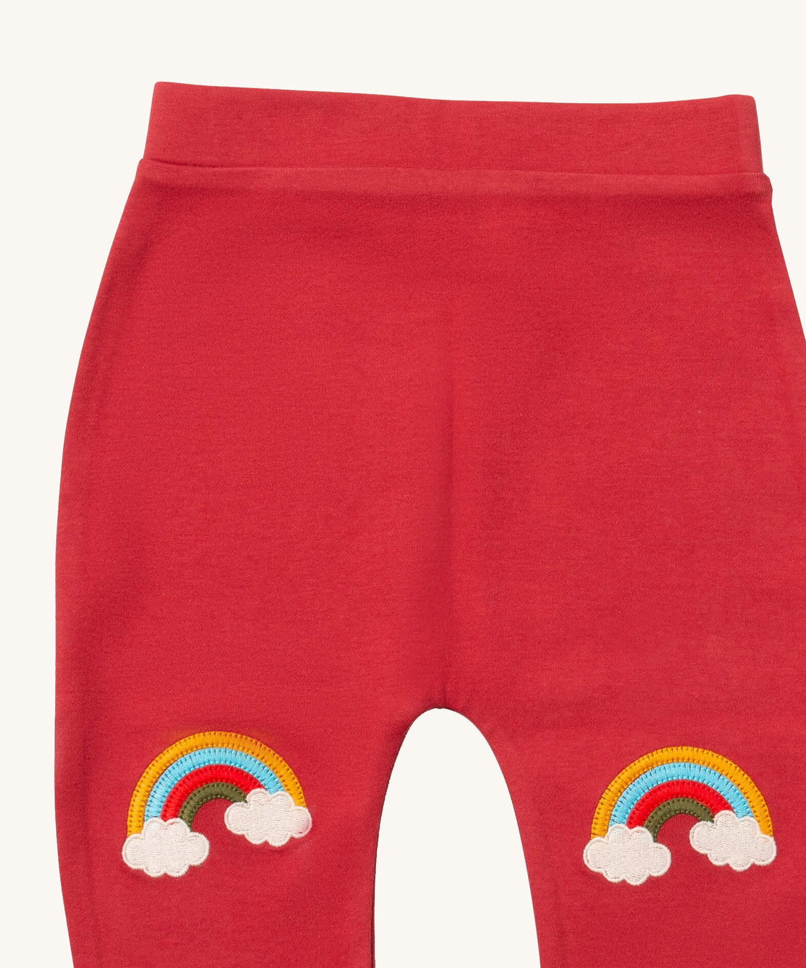 A close up of the Little Green Radicals soft red rainbow kneepatch joggers showing the rainbow embroidery kneepatch detail on pale red organic cotton fabric.