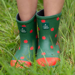A child wearing the Little Green Radicals kid wellington boots showing the length of the boots and the red apples print on the natural rubber material. Features a brand patch on the front of the wellington.