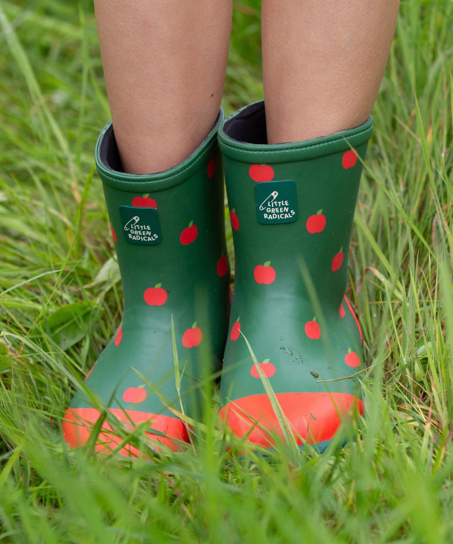 A child wearing the Little Green Radicals kid wellington boots showing the length of the boots and the red apples print on the natural rubber material. Features a brand patch on the front of the wellington.