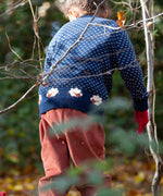 A child standing in woodland and wearing the Little Green Radicals navy sheep snuggle knitted jumper. Showing the white speckles and cute sheep continuing around the jumper