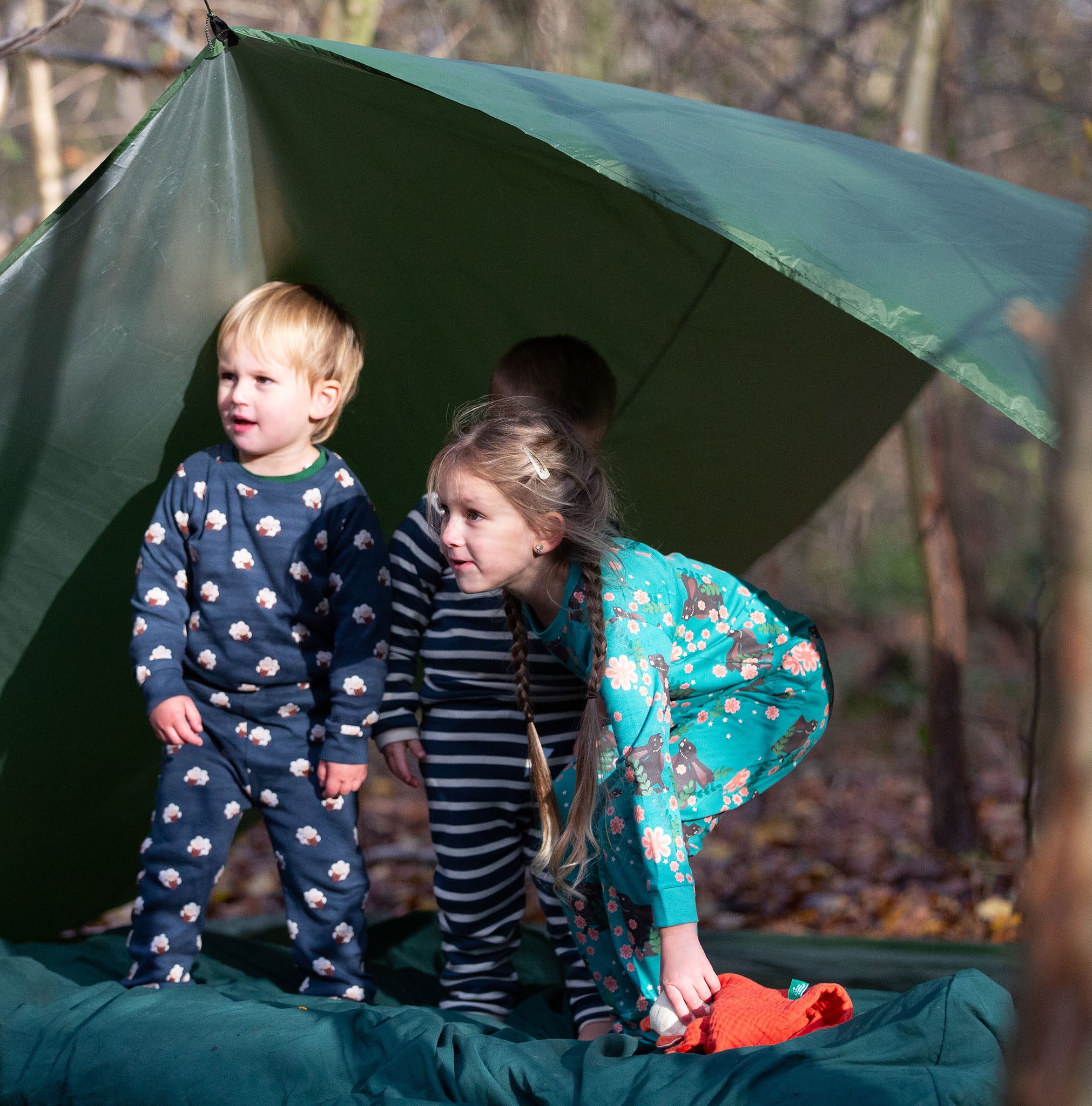 Children standing outside a tent in the forest. They are all wearing Little Green Radicals organic cotton pyjamas. One is wearing a pair with the Winter Rabbit on them, one in wearing navy and white striped design and the third is wearing the sleeping sheep print. Little green Radicals have a wide range of organic cotton, Fairtrade children's clothing an outerwear available here at Babipur.