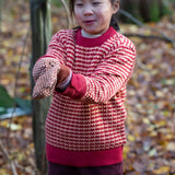 A child standing outside wearing the Little Green Radicals snuggly knitted jumper in juniper stripe available at Babipur.. A pale yellow and red striped GOTS organic jumper with a ribbed hem in pale red for a snug fit 