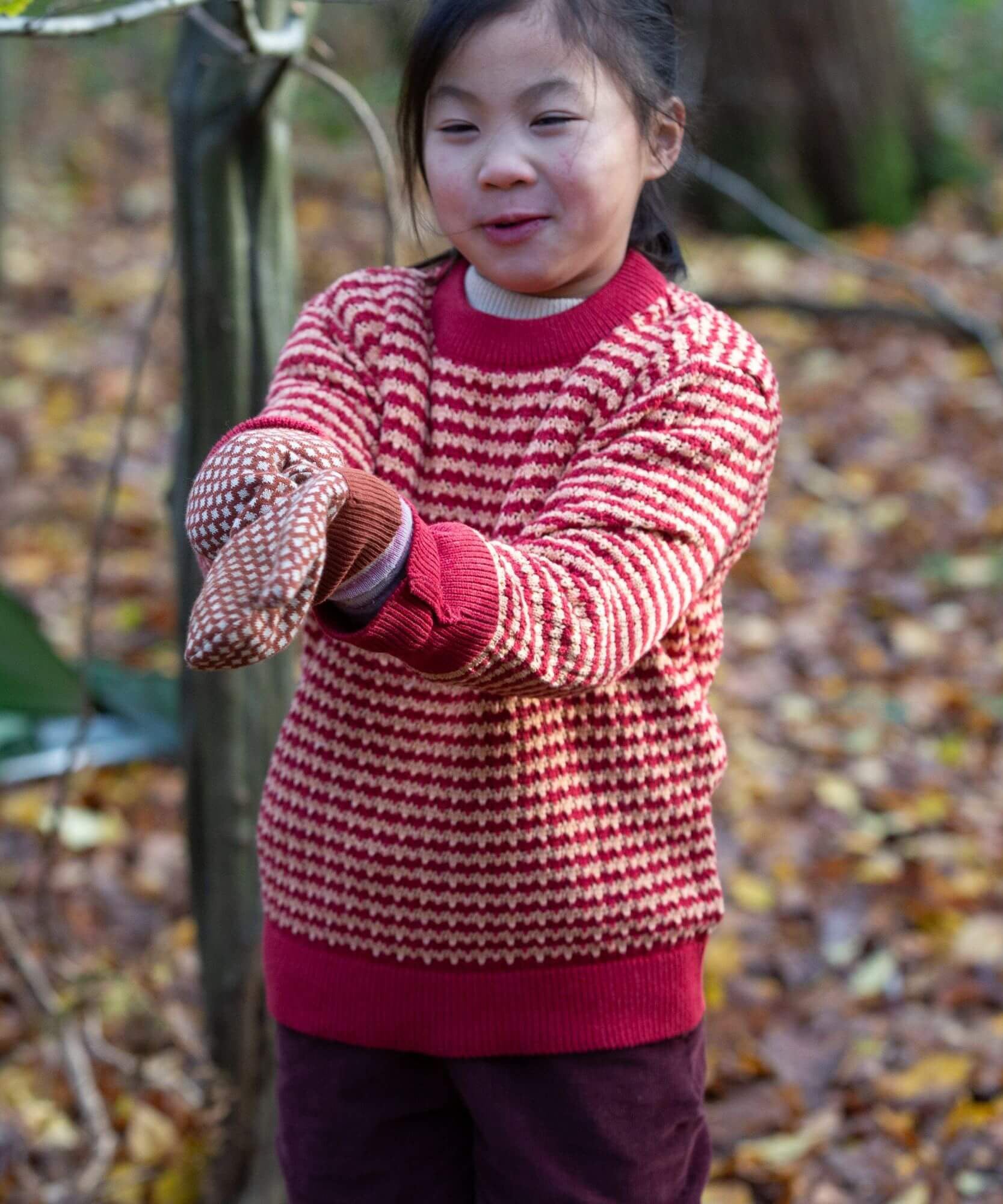 A child standing outside wearing the Little Green Radicals snuggly knitted jumper in juniper stripe available at Babipur.. A pale yellow and red striped GOTS organic jumper with a ribbed hem in pale red for a snug fit 
