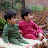 2 children sitting on a fluffy blanket wearing the holly berry pink and green chive waffle playsuit. A long sleeves and footless all in one suit made from GOTS organic cotton fabric with button fasteners and a small pouch chest pocket