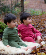 2 children sitting on a fluffy blanket wearing the holly berry pink and green chive waffle playsuit. A long sleeves and footless all in one suit made from GOTS organic cotton fabric with button fasteners and a small pouch chest pocket