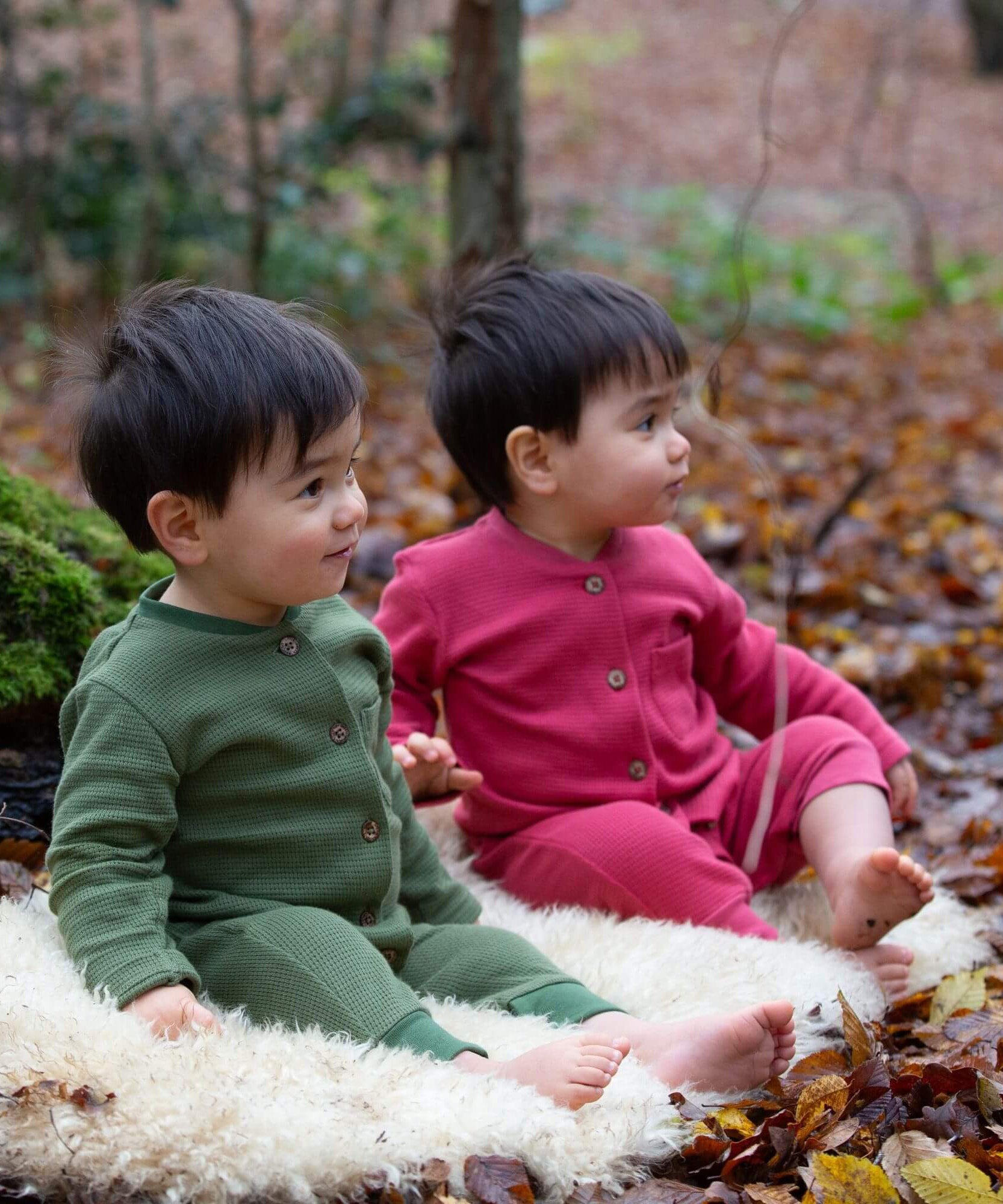 2 children sitting on a fluffy blanket wearing the holly berry pink and green chive waffle playsuit. A long sleeves and footless all in one suit made from GOTS organic cotton fabric with button fasteners and a small pouch chest pocket