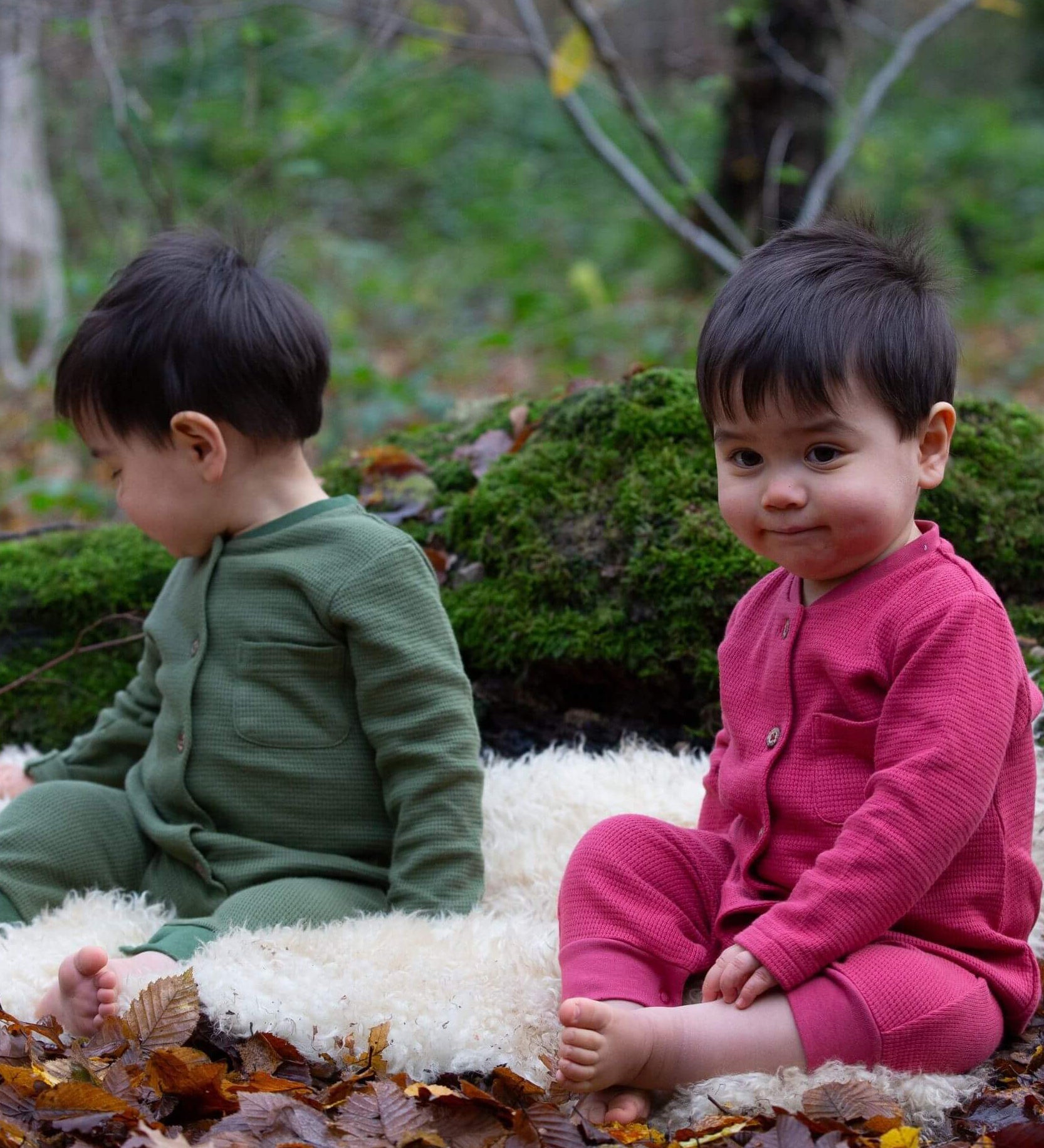 2 children wearing the holly berry pink and green chive waffle playsuit. A long sleeves all in one suit made from GOTs organic cotton fabric with button fasteners and a small pouch chest pocket
