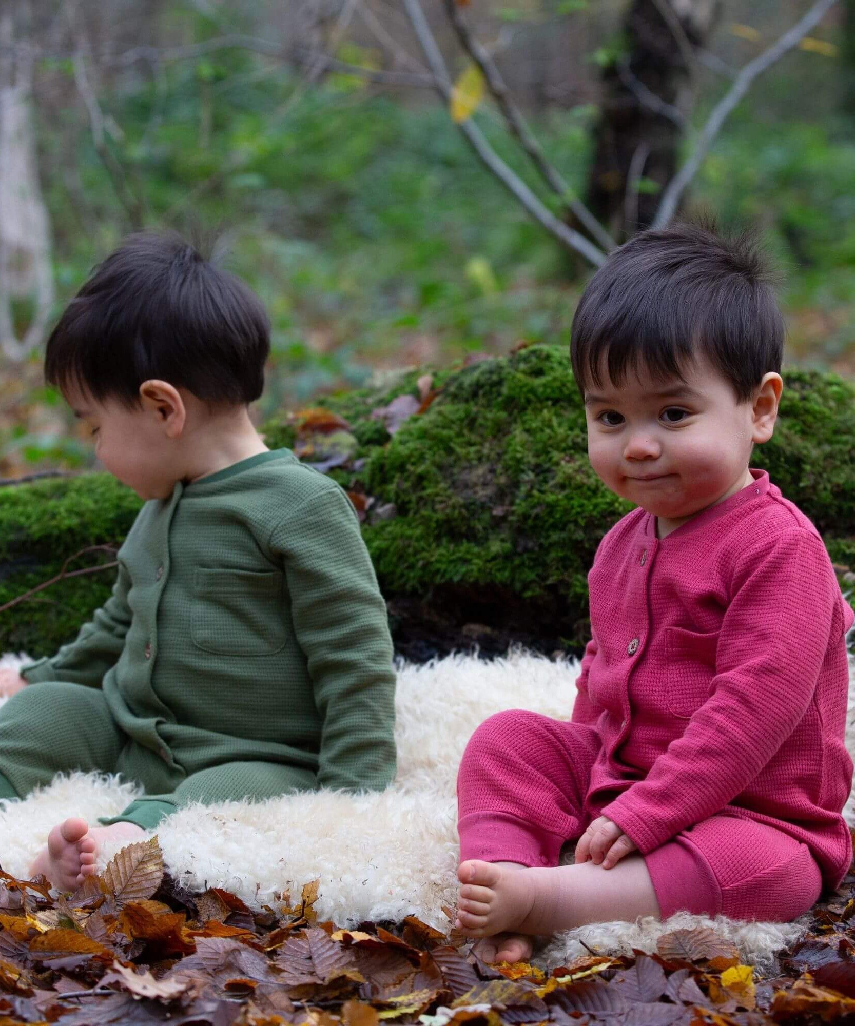 2 children wearing the holly berry pink and green chive waffle playsuit. A long sleeves all in one suit made from GOTs organic cotton fabric with button fasteners and a small pouch chest pocket