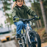 Child wearing a green helmet and riding Early Rider's Belter 16 inch wheel children's bike on a gravel path with a forest background