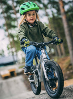 Child wearing a green helmet and riding Early Rider's Belter 16 inch wheel children's bike on a gravel path with a forest background