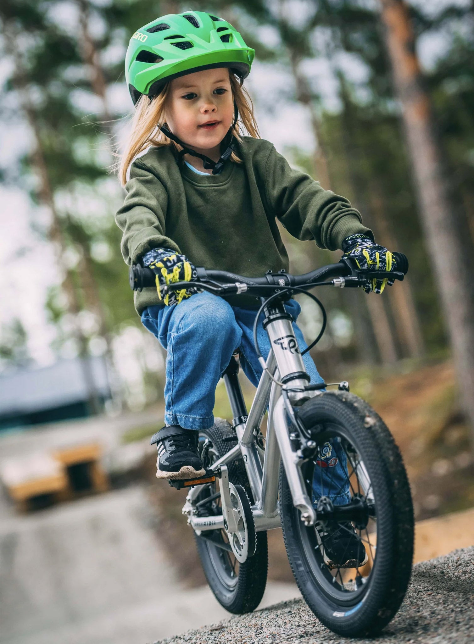 Child wearing a green helmet and riding Early Rider's Belter 16 inch wheel children's bike on a gravel path with a forest background