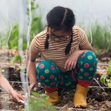 Child wearing the Little Green Radicals apple picking green comfy organic cotton joggers with a short sleeved gold and cream striped t-shirt and yellow wellington boots whilst gardening. 