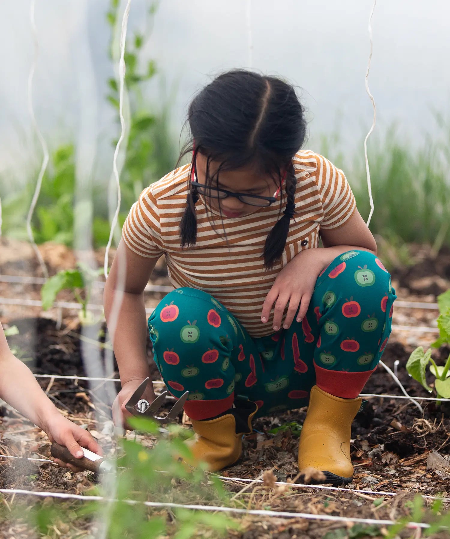 Child wearing the Little Green Radicals apple picking green comfy organic cotton joggers with a short sleeved gold and cream striped t-shirt and yellow wellington boots whilst gardening. 