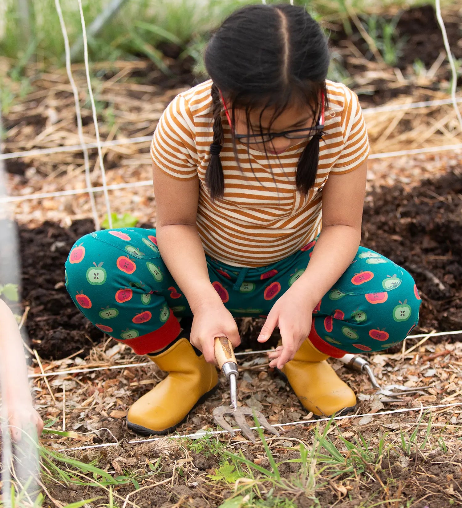 Child wearing the Little Green Radicals apple picking green comfy organic cotton joggers with a short sleeved gold and cream striped t-shirt and yellow wellington boots. 