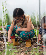 Child wearing the Little Green Radicals apple picking green comfy organic cotton joggers with a short sleeved gold and cream striped t-shirt and yellow wellington boots whilst gardening. 