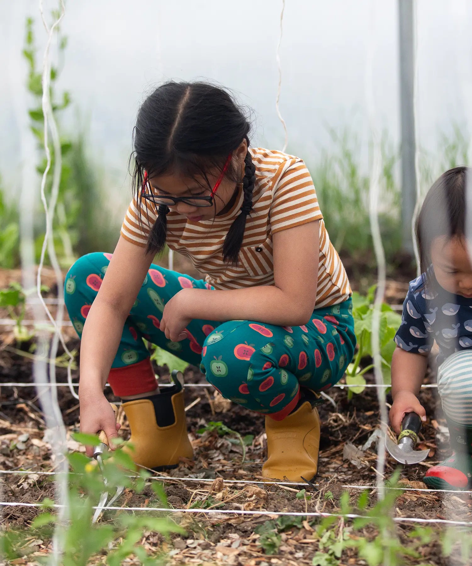 Child wearing the Little Green Radicals apple picking green comfy organic cotton joggers with a short sleeved gold and cream striped t-shirt and yellow wellington boots whilst gardening. 