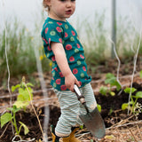 Child wearing the Little Green Radicals apple picking organic cotton playset outfit with yellow wellington boots. A short sleeved t-shirt with red and green repeat apple print on a green background with green and cream striped joggers. 