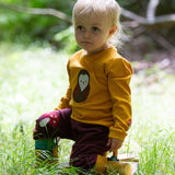 A child wearing the Little Green Radicals Applique Long Sleeve yellow gold coloured Top with Brown Owl applique on the front and a mushroom on the sleeve. This top is part of a range of organic cotton children's clothing available at Babipur.