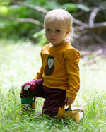 A child wearing the Little Green Radicals Applique Long Sleeve yellow gold coloured Top with Brown Owl applique on the front and a mushroom on the sleeve. This top is part of a range of organic cotton children's clothing available at Babipur.