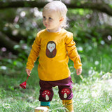 A child standing on grass wearing the Little Green Radicals Applique Long Sleeve yellow gold coloured Top with Brown Owl applique on the front and a mushroom on the sleeve with marron mushroom knee patch joggers. These are part of a range of organic cotton children's clothing available at Babipur.