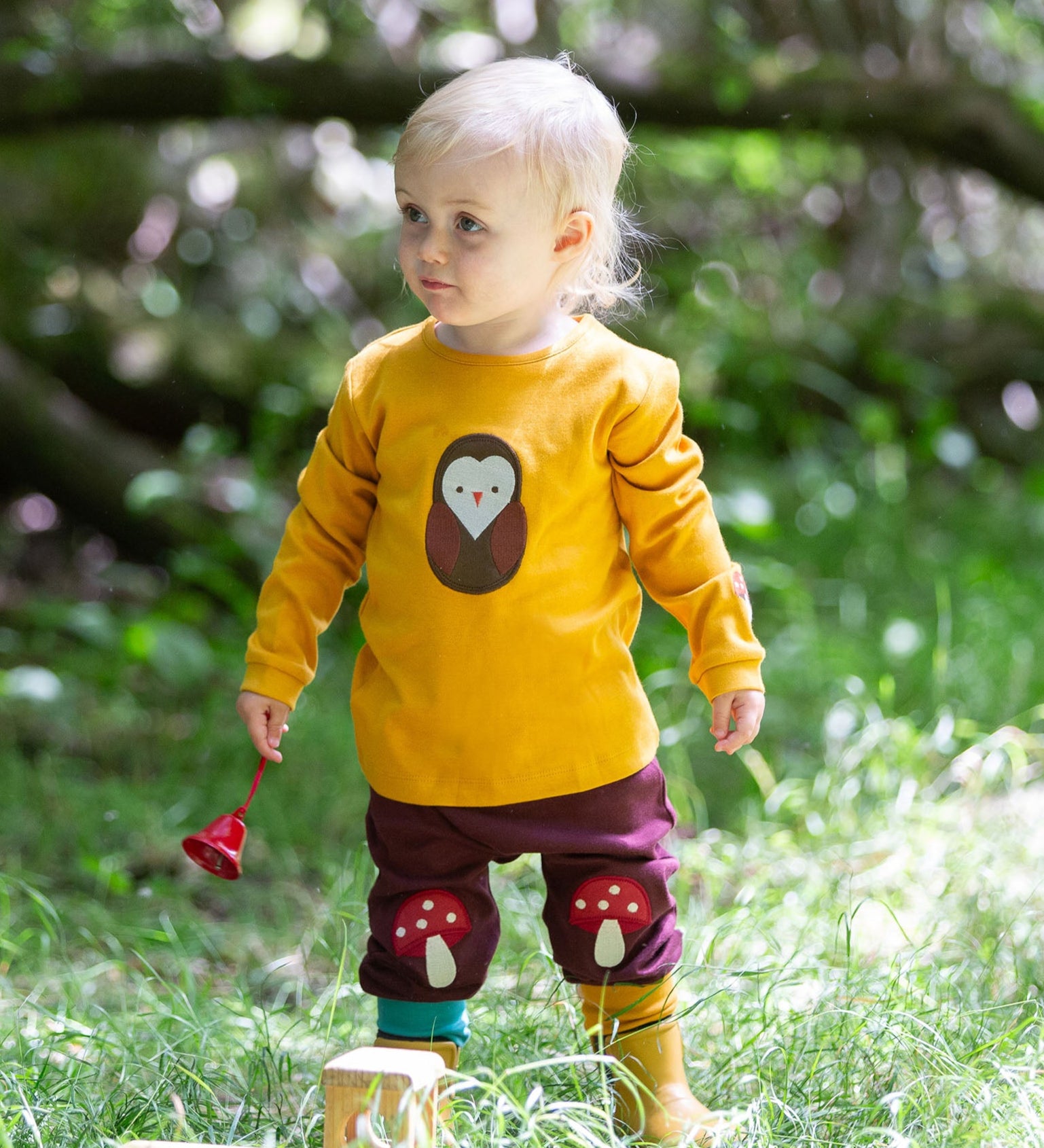 A child standing on grass wearing the Little Green Radicals Applique Long Sleeve yellow gold coloured Top with Brown Owl applique on the front and a mushroom on the sleeve with marron mushroom knee patch joggers. These are part of a range of organic cotton children's clothing available at Babipur.