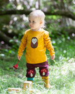 A child standing on grass wearing the Little Green Radicals Applique Long Sleeve yellow gold coloured Top with Brown Owl applique on the front and a mushroom on the sleeve with marron mushroom knee patch joggers. These are part of a range of organic cotton children's clothing available at Babipur.