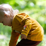 A child wearing the Little Green Radicals Applique Long Sleeve yellow gold coloured Top with Brown Owl applique on the front and a mushroom on the sleeve. This top is part of a range of organic cotton children's clothing available at Babipur.