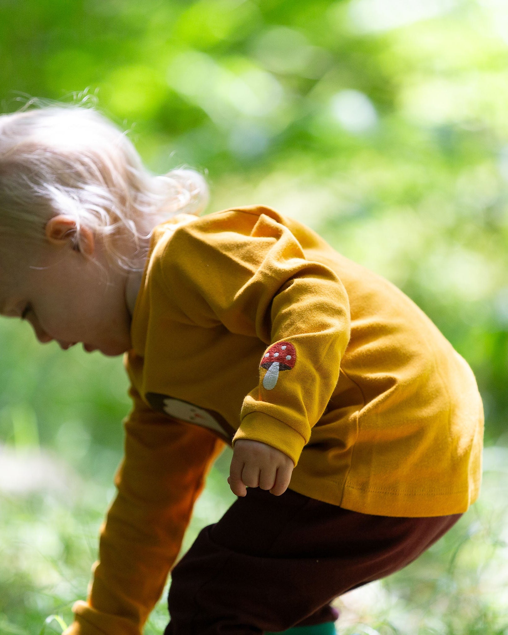 A child wearing the Little Green Radicals Applique Long Sleeve yellow gold coloured Top with Brown Owl applique on the front and a mushroom on the sleeve. This top is part of a range of organic cotton children's clothing available at Babipur.